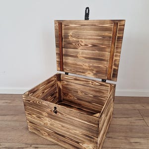 May include: A wooden chest with a hinged lid, open to reveal the interior. The chest has a rustic, burnt wood finish with visible wood grain. A black metal latch secures the lid. The chest is sitting on a wooden floor.