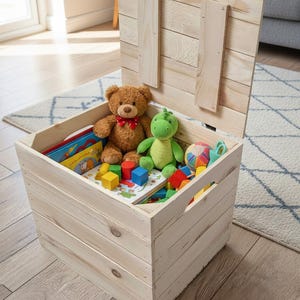 May include: A light-colored wooden toy chest with the lid open, revealing a teddy bear, a green stuffed dinosaur, colorful blocks, and books. The chest is sitting on a light-colored wooden floor.