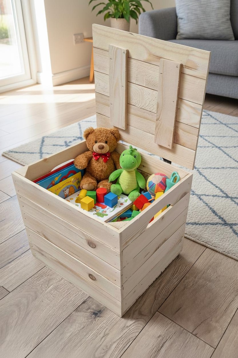 May include: A light-colored wooden toy chest with the lid open, revealing a teddy bear, a green stuffed dinosaur, colorful blocks, and books. The chest is sitting on a light-colored wooden floor.