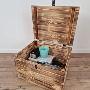 K&ouml;nnte beinhalten: Eine rustikale Holz-Truhe mit Scharnierdeckel, in Braunt&ouml;nen gebeizt. Die Truhe ist ge&ouml;ffnet und zeigt verschiedene Gegenst&auml;nde im Inneren, darunter einen kleinen blauen Eimer. Die Holzmaserung ist sichtbar und tr&auml;gt zur nat&uuml;rlichen &Auml;sthetik der Truhe bei.