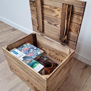 May include: A rustic wooden storage chest with a hinged lid, open to reveal its contents. Inside are several boxes, including a puzzle box with a floral design. The chest is made of light brown wood with darker burn marks.