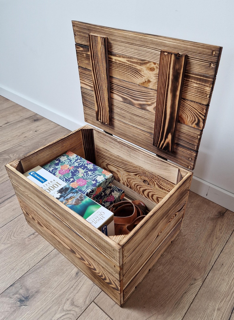 May include: A rustic wooden storage chest with a hinged lid, open to reveal its contents. Inside are several boxes, including a puzzle box with a floral design. The chest is made of light brown wood with darker burn marks.