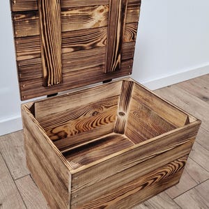 May include: A rustic wooden storage box with a hinged lid, crafted from light brown wood with darker, charred accents. The box is open, revealing its interior, and sits on a wooden floor.