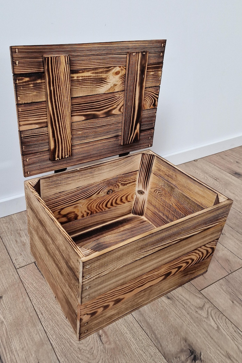May include: A rustic wooden storage box with a hinged lid, crafted from light brown wood with darker, charred accents. The box is open, revealing its interior, and sits on a wooden floor.