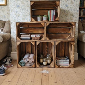 Peut inclure: Étagère composée de six caisses en bois marron foncé. Les caisses contiennent des livres, des plantes, des serviettes, des bocaux et des magazines. L'étagère est placée contre un papier peint à motifs floraux.