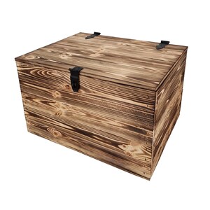 K&ouml;nnte beinhalten: Eine quadratische Holz-Aufbewahrungsbox mit rustikalem, verbranntem Holzfinish. Die Truhe hat einen Klappdeckel mit einem schwarzen Metallverschluss und zwei kleineren Metallscharnieren. Die Holzmaserung ist sichtbar.