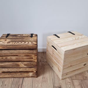 May include: Two wooden storage chests. One is stained a dark brown, the other is natural wood. Both have hinged lids with black metal hardware. The chests are constructed from horizontal wooden planks and are suitable for various storage needs.