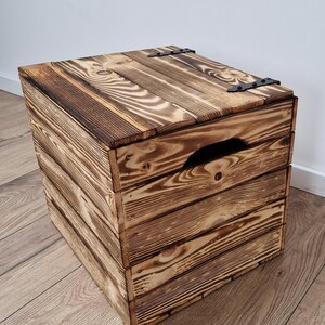 May include: A rustic wooden storage box with a hinged lid and a handle cut into the front. The box is made of stained wood with visible grain and dark metal hinges. The box is square and sits on a wooden floor.