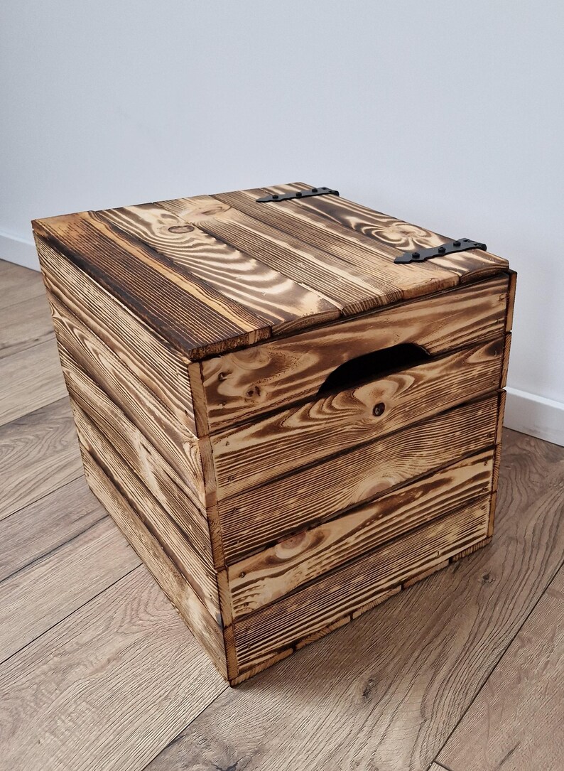 May include: A rustic wooden storage box with a hinged lid and a handle cut into the front. The box is made of stained wood with visible grain and dark metal hinges. The box is square and sits on a wooden floor.