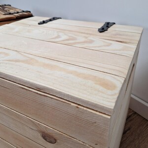 May include: A light-colored wooden chest with a hinged lid. The chest is constructed from planks of wood and features black metal hinges. The wood grain is visible, and the chest appears to be new.