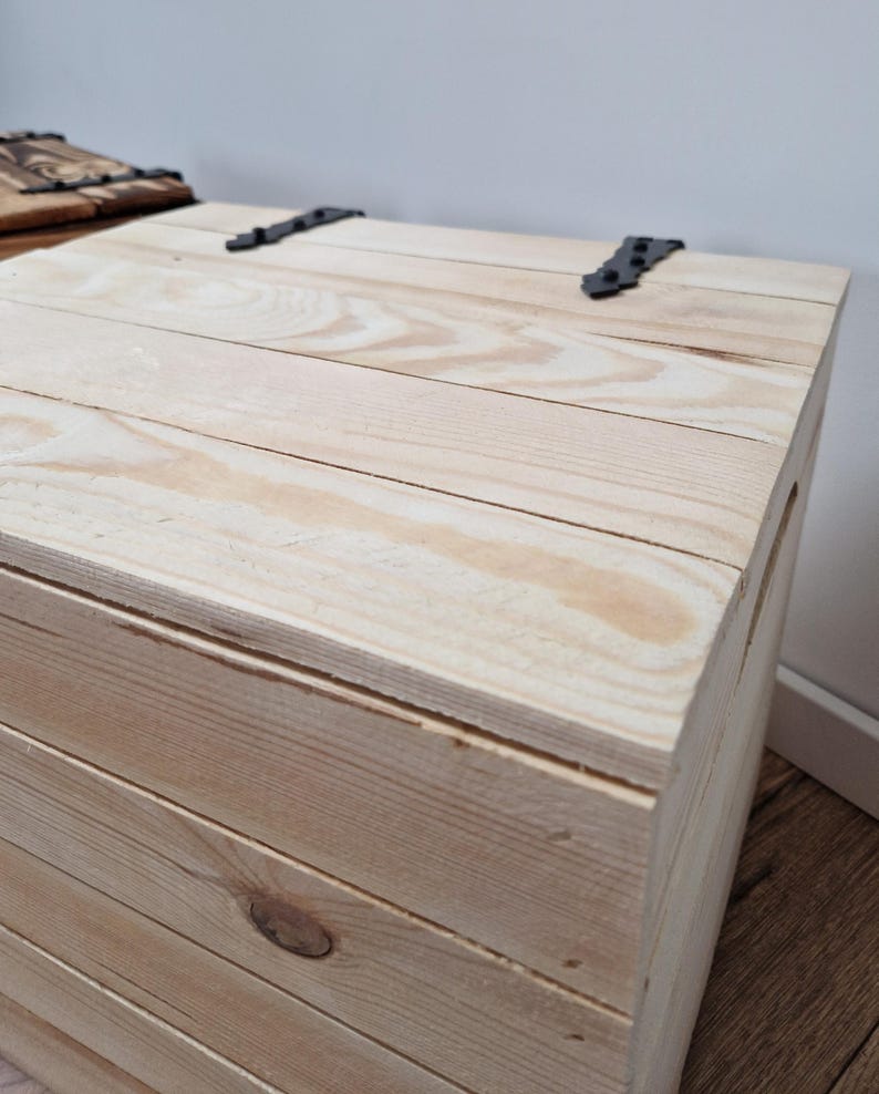 May include: A light-colored wooden chest with a hinged lid. The chest is constructed from planks of wood and features black metal hinges. The wood grain is visible, and the chest appears to be new.