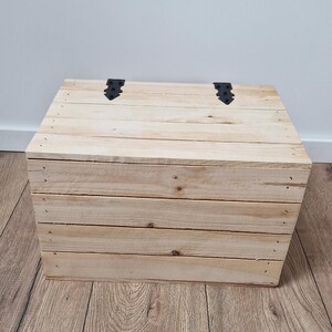 May include: A wooden chest made of light-colored planks, with a hinged lid and black metal hardware. The chest is rectangular and appears to be handmade, with visible nail heads. The overall design is rustic and simple.