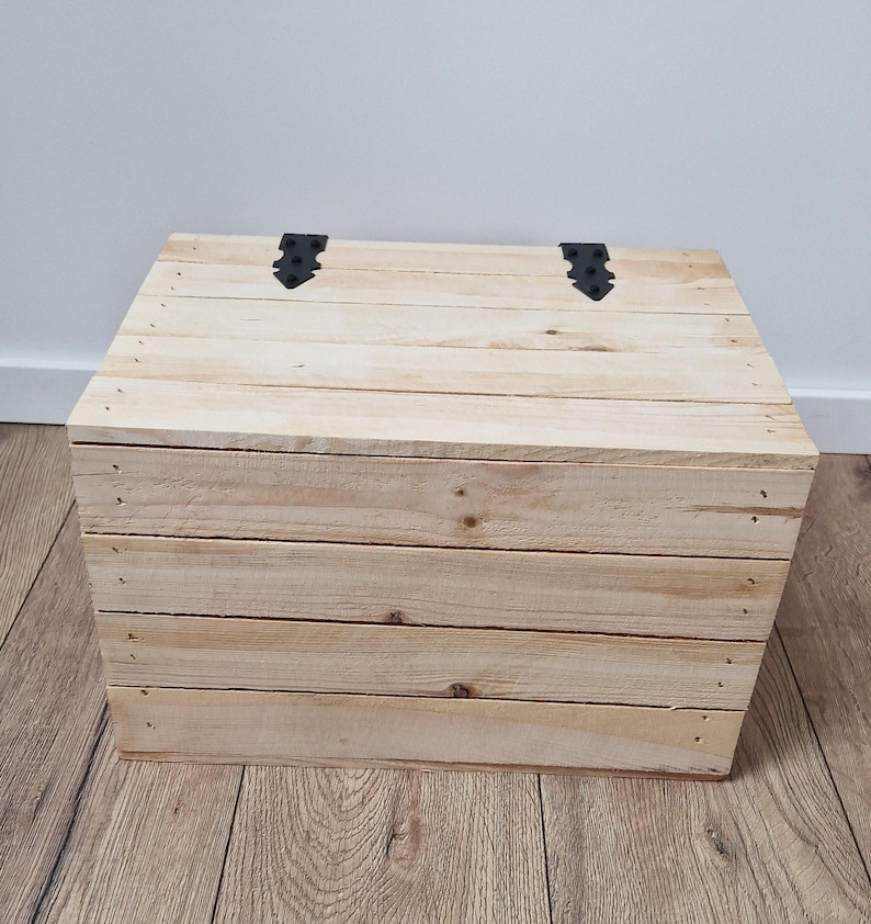 May include: A wooden chest made of light-colored planks, with a hinged lid and black metal hardware. The chest is rectangular and appears to be handmade, with visible nail heads. The overall design is rustic and simple.