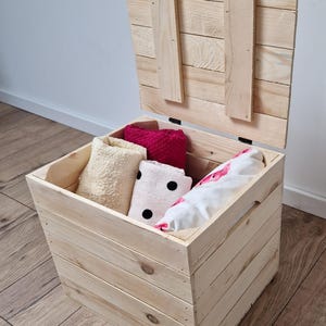 May include: A light-colored wooden storage chest with the lid slightly ajar, revealing folded towels in cream, pink, and white with black polka dots. The chest is made of unfinished wood and sits on a wooden floor.