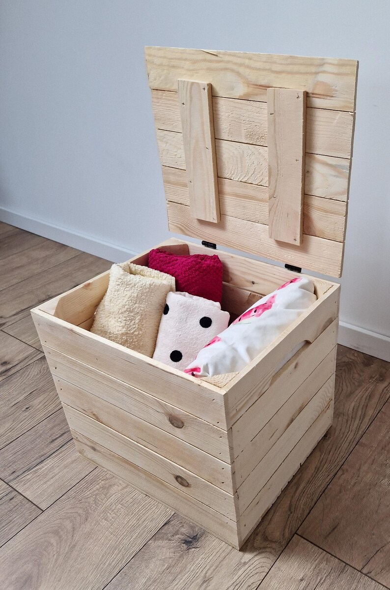 May include: A light-colored wooden storage chest with the lid slightly ajar, revealing folded towels in cream, pink, and white with black polka dots. The chest is made of unfinished wood and sits on a wooden floor.