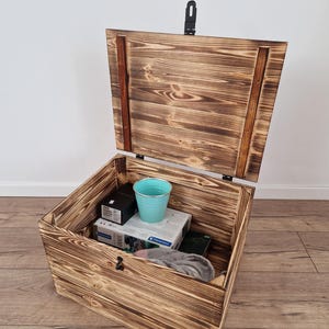 May include: Open wooden storage chest with a hinged lid and a black latch. The chest is made of light brown wood with a visible grain pattern. Inside the chest are various items, including a blue bucket and boxes.