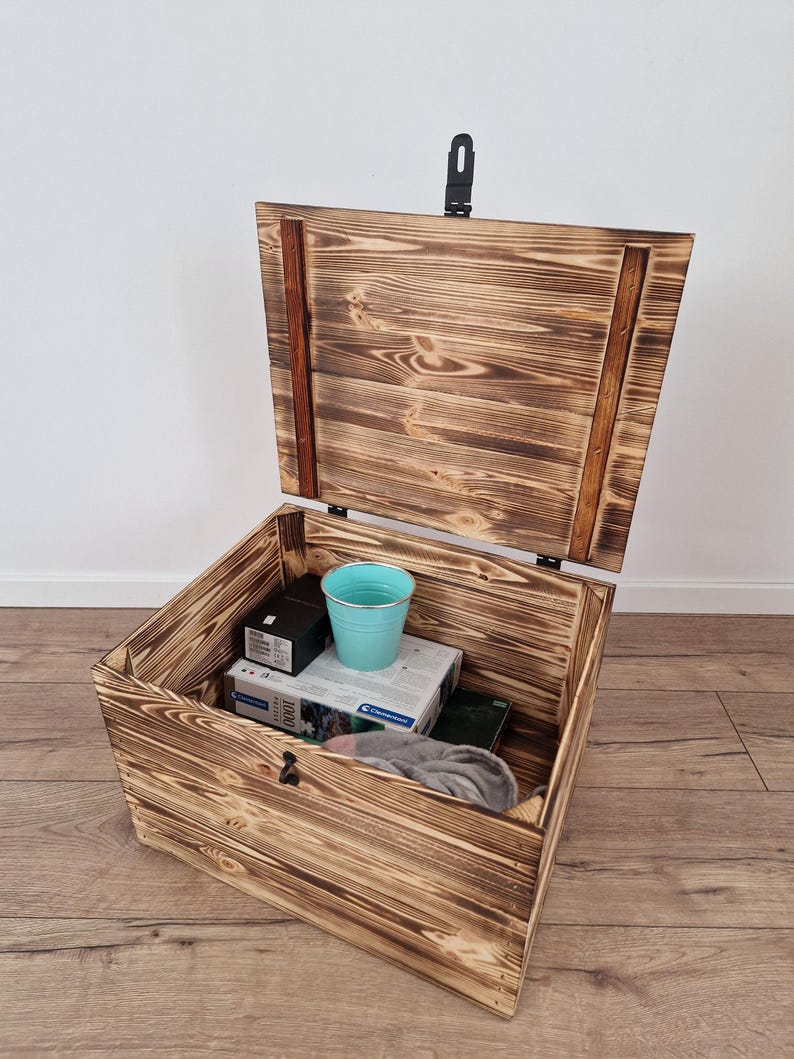 May include: Open wooden storage chest with a hinged lid and a black latch. The chest is made of light brown wood with a visible grain pattern. Inside the chest are various items, including a blue bucket and boxes.