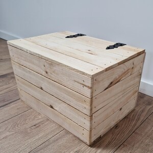 May include: A light-colored wooden storage box with a hinged lid and black metal hardware. The box is constructed from horizontal wooden planks and is suitable for various storage needs.