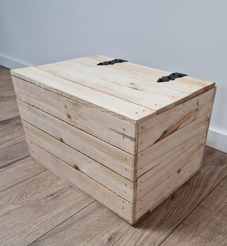 May include: A light-colored wooden storage box with a hinged lid and black metal hardware. The box is constructed from horizontal wooden planks and is suitable for various storage needs.