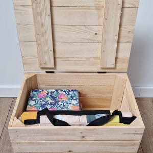 May include: A wooden storage chest with a hinged lid, open to reveal a floral patterned fabric and a black handled bag. The chest is made of light-colored wood and sits on a wooden floor.