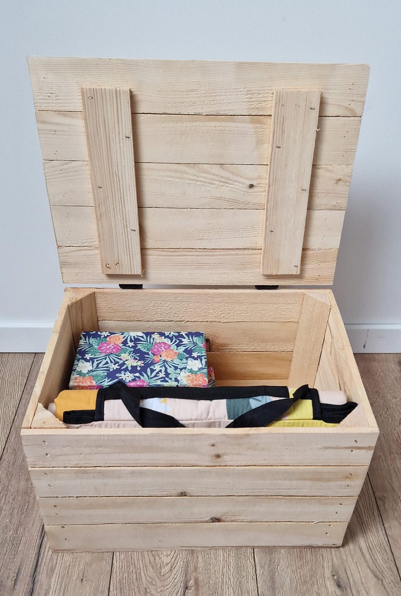 May include: A wooden storage chest with a hinged lid, open to reveal a floral patterned fabric and a black handled bag. The chest is made of light-colored wood and sits on a wooden floor.