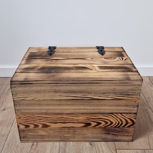 May include: A rustic wooden chest with a dark, burnt finish. The chest is rectangular with a hinged lid and black metal hinges. The wood grain is visible, adding to the vintage aesthetic. The chest is a storage solution.