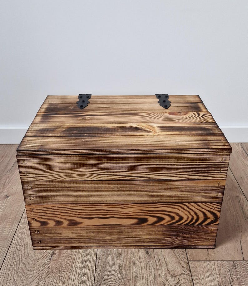 May include: A rustic wooden chest with a dark, burnt finish. The chest is rectangular with a hinged lid and black metal hinges. The wood grain is visible, adding to the vintage aesthetic. The chest is a storage solution.