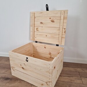 K&ouml;nnte beinhalten: Eine hellfarbene Holz-Truhe mit teilweise ge&ouml;ffnetem Deckel. Die Truhe ist aus Holzplanken gefertigt, mit sichtbaren Knoten und Maserungen. Sie hat einen schwarzen Verschluss und Scharniere. Die Truhe steht auf einem Holzboden.
