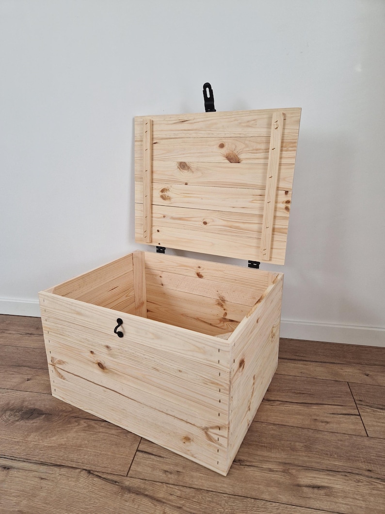 K&ouml;nnte beinhalten: Eine hellfarbene Holz-Truhe mit teilweise ge&ouml;ffnetem Deckel. Die Truhe ist aus Holzplanken gefertigt, mit sichtbaren Knoten und Maserungen. Sie hat einen schwarzen Verschluss und Scharniere. Die Truhe steht auf einem Holzboden.