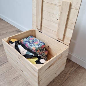 May include: Open wooden storage chest with a hinged lid. The chest is made of light-colored wood and contains a floral-patterned box and other items. The lid is propped open, revealing the contents inside. The chest is sitting on a wooden floor.