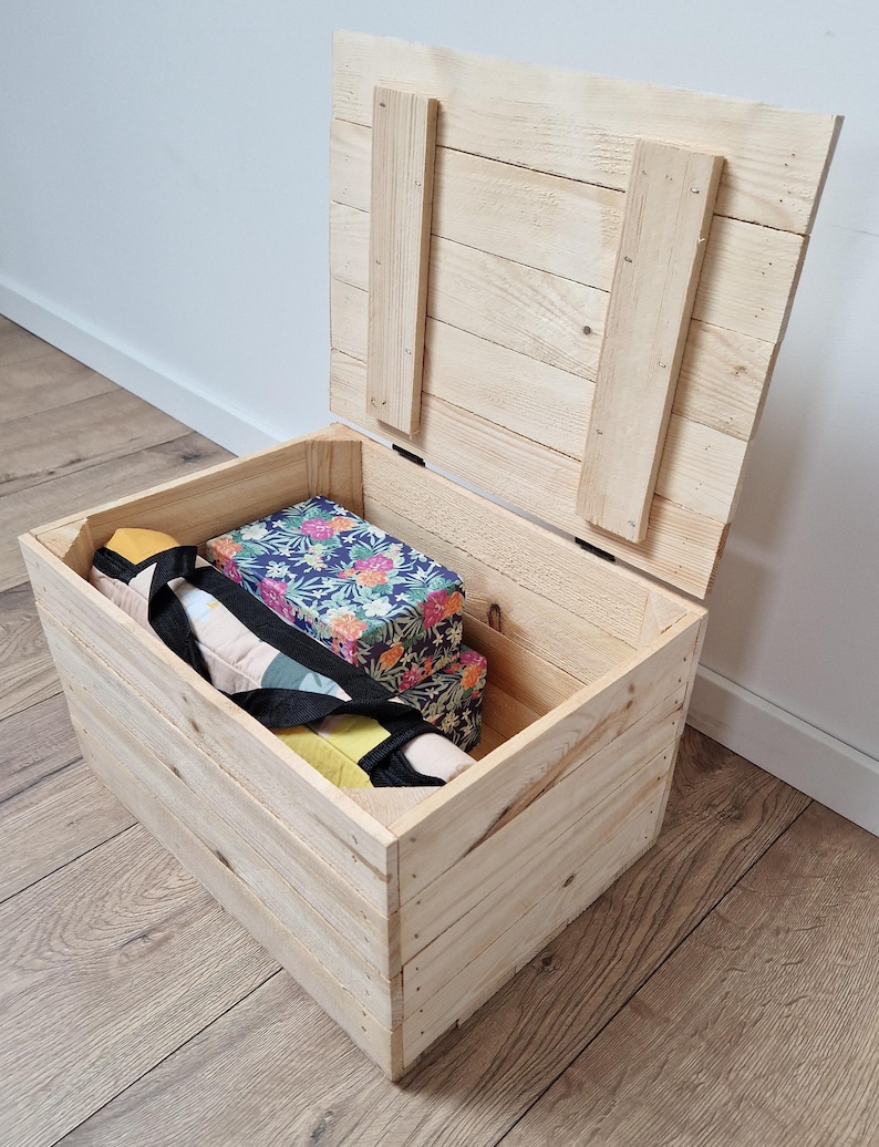 May include: Open wooden storage chest with a hinged lid. The chest is made of light-colored wood and contains a floral-patterned box and other items. The lid is propped open, revealing the contents inside. The chest is sitting on a wooden floor.