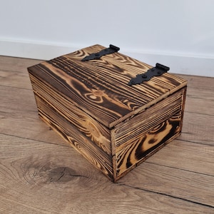 May include: A rustic wooden box with a hinged lid and black metal hardware. The box is made of stained wood with a visible grain pattern. The box is rectangular and appears to be handmade.