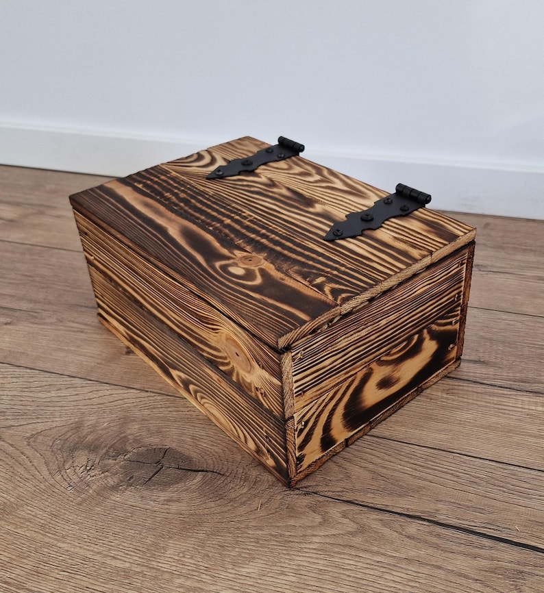 May include: A rustic wooden box with a hinged lid and black metal hardware. The box is made of stained wood with a visible grain pattern. The box is rectangular and appears to be handmade.