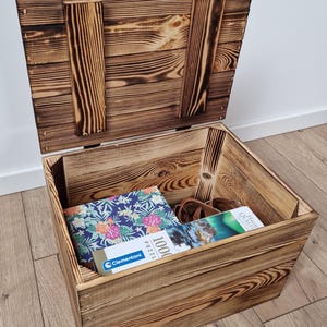 May include: A rustic wooden storage chest with a hinged lid, stained in a warm brown tone. The chest is open, revealing a puzzle box with a floral design, a pair of sunglasses, and another puzzle box. The chest is sitting on a wooden floor.