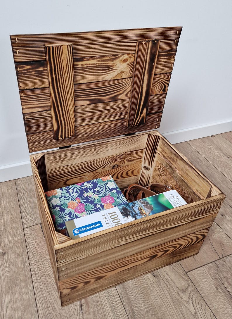May include: A rustic wooden storage chest with a hinged lid, stained in a warm brown tone. The chest is open, revealing a puzzle box with a floral design, a pair of sunglasses, and another puzzle box. The chest is sitting on a wooden floor.