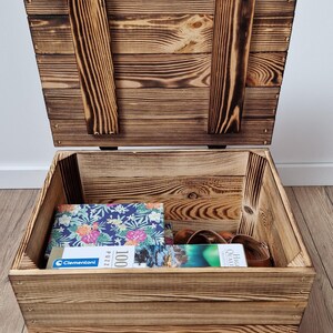 May include: Open wooden storage chest with a hinged lid and two wooden support beams. The chest is filled with a puzzle box, a belt, and other items. The wood has a natural grain and a warm, brown color.