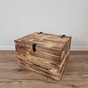 K&ouml;nnte beinhalten: Eine quadratische Holzkiste mit Scharnierdeckel und schwarzen Metallbeschl&auml;gen. Die Kiste hat eine rustikale, verbrannte Holzoberfl&auml;che, die die nat&uuml;rliche Holzmaserung zeigt. Die Kiste steht auf einem Holzboden.
