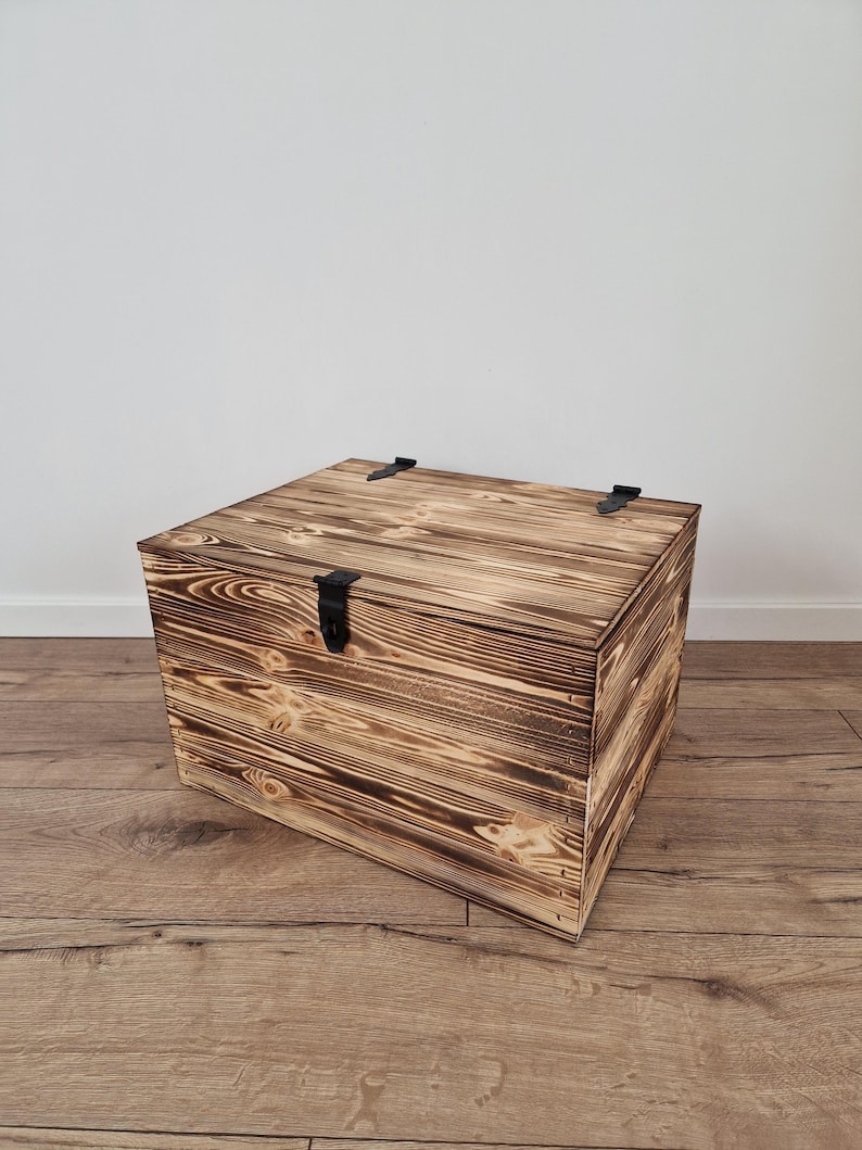 K&ouml;nnte beinhalten: Eine quadratische Holzkiste mit Scharnierdeckel und schwarzen Metallbeschl&auml;gen. Die Kiste hat eine rustikale, verbrannte Holzoberfl&auml;che, die die nat&uuml;rliche Holzmaserung zeigt. Die Kiste steht auf einem Holzboden.