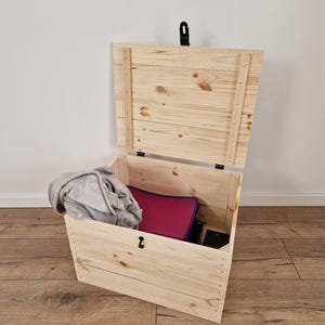 May include: A wooden storage chest with the lid partially open, revealing a gray blanket, a magenta folder, and a black object inside. The chest is made of light-colored wood and has a black latch.