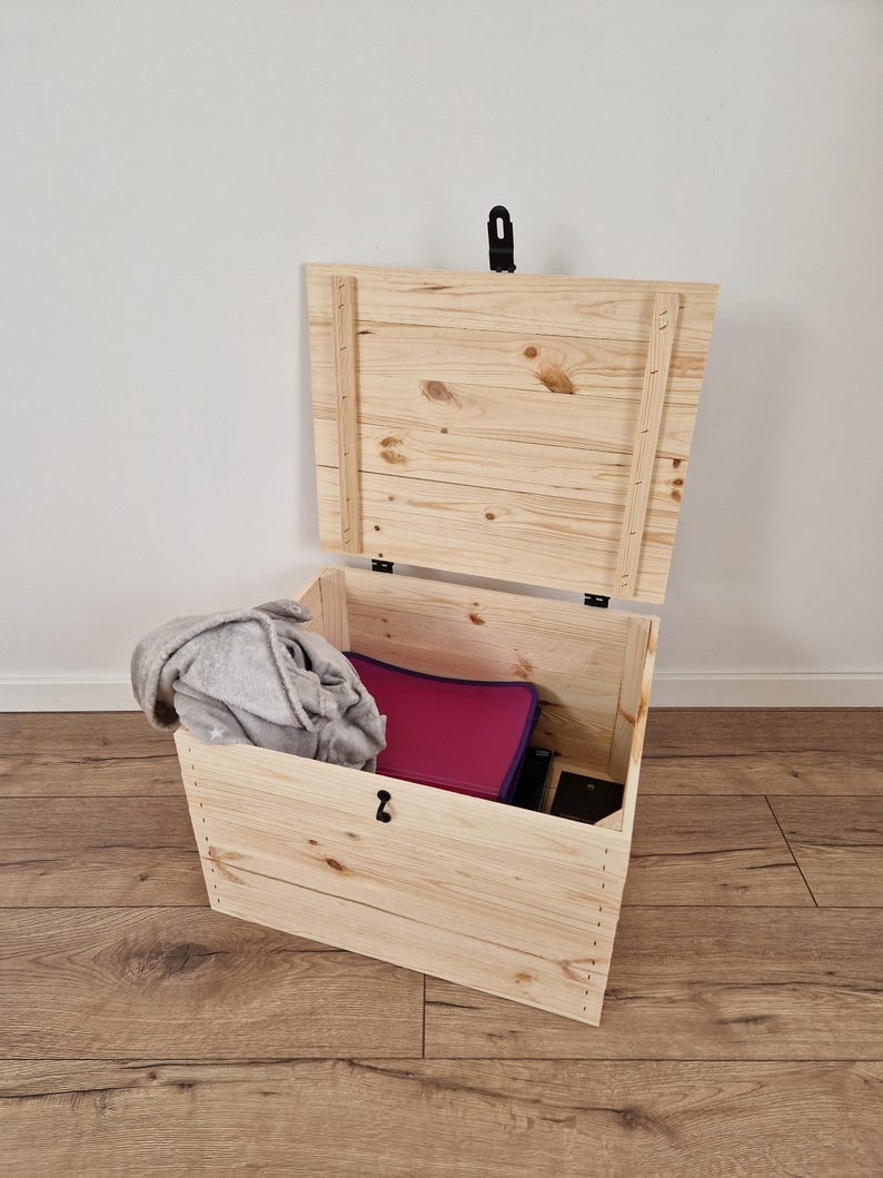 May include: A wooden storage chest with the lid partially open, revealing a gray blanket, a magenta folder, and a black object inside. The chest is made of light-colored wood and has a black latch.