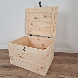 May include: A light-colored wooden storage chest with the lid partially open. The chest is rectangular and constructed from planks of wood, with visible wood grain. The lid is attached with hinges and has a black handle. The chest is on a wooden floor.