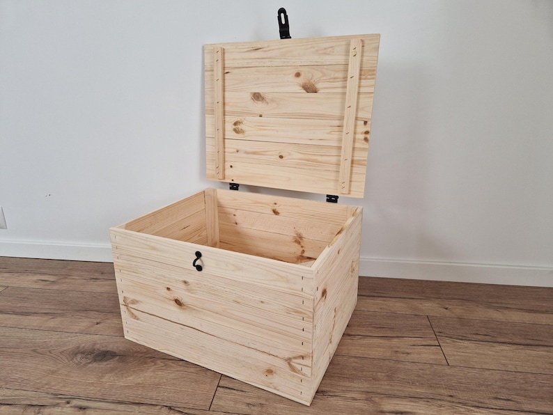 May include: A light-colored wooden storage chest with the lid partially open. The chest is rectangular and constructed from planks of wood, with visible wood grain. The lid is attached with hinges and has a black handle. The chest is on a wooden floor.