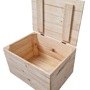 May include: An open, light-colored wooden storage chest with a hinged lid. The chest is constructed from planks of wood, with the lid secured by two vertical wooden supports. The interior of the chest is visible, showcasing the natural wood grain.