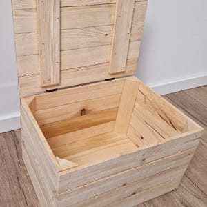 May include: An open, unfinished wooden storage chest with a hinged lid. The chest is constructed from light-colored wooden planks, with the lid propped open. The chest is rectangular and appears to be handmade.