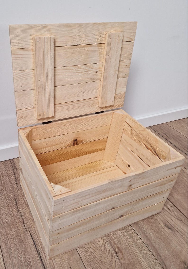 May include: An open, unfinished wooden storage chest with a hinged lid. The chest is constructed from light-colored wooden planks, with the lid propped open. The chest is rectangular and appears to be handmade.