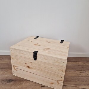 May include: A light-colored wooden storage chest with a rectangular shape. The chest has a hinged lid and is secured with a black metal latch. The wood grain is visible, and the chest is sitting on a wooden floor.