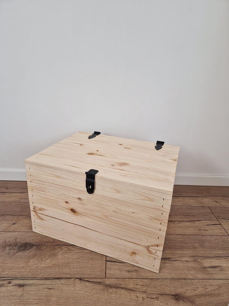 May include: A light-colored wooden storage chest with a rectangular shape. The chest has a hinged lid and is secured with a black metal latch. The wood grain is visible, and the chest is sitting on a wooden floor.