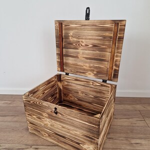 K&ouml;nnte beinhalten: Eine Holzkiste mit einem Scharnierdeckel, der ge&ouml;ffnet ist, um das Innere zu zeigen. Die Kiste hat eine rustikale, verbrannte Holzoberfl&auml;che mit sichtbarer Holzmaserung. Ein schwarzer Metallverschluss sichert den Deckel. Die Kiste steht auf einem Holzboden.