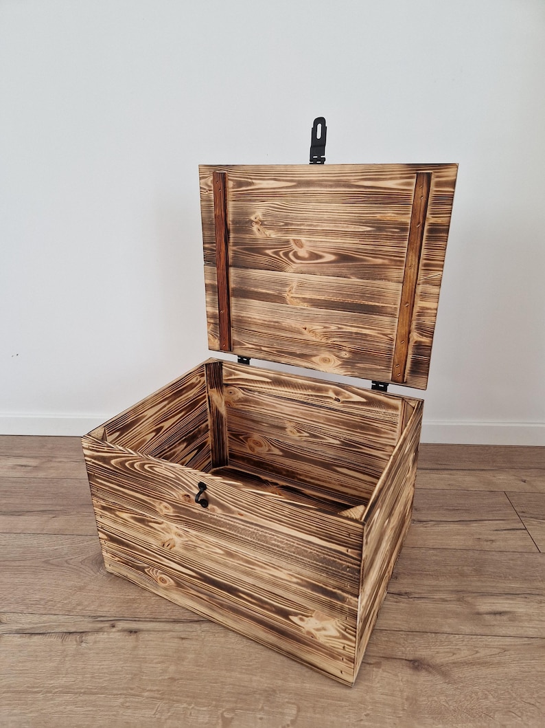 K&ouml;nnte beinhalten: Eine Holzkiste mit einem Scharnierdeckel, der ge&ouml;ffnet ist, um das Innere zu zeigen. Die Kiste hat eine rustikale, verbrannte Holzoberfl&auml;che mit sichtbarer Holzmaserung. Ein schwarzer Metallverschluss sichert den Deckel. Die Kiste steht auf einem Holzboden.