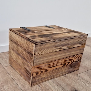May include: A rustic wooden storage box with a hinged lid. The box is constructed from light brown wood with visible grain patterns and dark brown burn marks. Black metal hinges secure the lid. The box is square-shaped and appears to be handmade.
